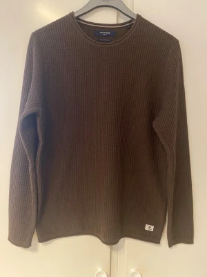 Brun stickad tröja Jack & Jones - Mörkbrun stickad tröja från Jack & Jones med rund halsringning och lång ärm. Tröjan har en grovstickad struktur och en liten logolapp nertill. Perfekt för kyliga dagar och enkel att matcha med olika outfits.