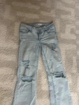Ljusblå slitna bootcut jeans - Säljer ett par ljusblå jeans från Nelly med bootcut-modell och flera slitningar och hål på både fram- och baksida. Jeansen har egengjorda slitningar och hål. Det är slitningar längst ner på jeansen men de syns inte när man har på sig dem. Original pris 700.