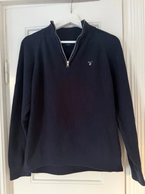 Mörkblå stickad tröja från Gant - Snygg mörkblå stickad tröja från Gant med halv dragkedja och diskret logga på bröstet. Tröjan har ribbad struktur, hög krage och långa ärmar. Perfekt för lager på lager och en klassisk look.