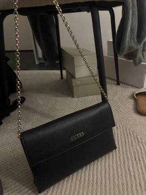 Svart väska från Guess - Svart axelväska från Guess i slät, strukturerad finish. Smal och stilren modell med lockstängning och guldfärgad kedjerem. Praktisk storlek som rymmer mobil, kort och mindre nödvändigheter. En tidlös väska som funkar både till vardag och kväll.🖤