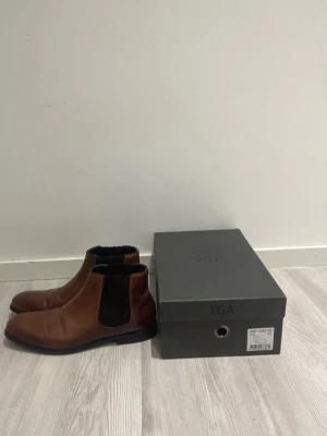 Bruna Chelsea boots från TGA - Snygga bruna Chelsea boots från TGA i slätt läder med elastiska svarta paneler på sidorna och låg klack. Klassisk siluett som funkar till många olika stilar. Skorna har en mörk sula och kommer med originalkartong.