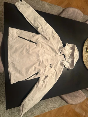 Vit vindjacka från Under Armour - Snygg vit/beige vindjacka från Under Armour med dragkedja framtill, två sidofickor med svarta dragkedjor och justerbar huva. Tillverkad i lätt och tåligt syntetmaterial, perfekt för blåsiga dagar. Diskret logga på bröstet och sportig passform.