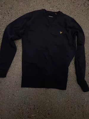 Svart sweatshirt från Lyle & Scott - Svart sweatshirt från Lyle & Scott med rund halsringning och klassisk gul logga broderad på bröstet. Tröjan har ribbade muddar vid ärmslut och nederkant. Perfekt för en stilren och avslappnad look. Köpte en storlek m men fick xs av misstag av en annan användare