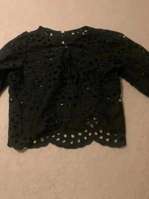 Svart broderad blus med hålmönster - Snygg svart blus med broderade detaljer och hålmönster över hela plagget. Blusen har lång ärm och knytning framtill vid halsen. Materialet känns som bomull och har en vågig kant nertill för extra stilpoäng.