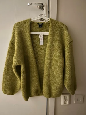  Ny Grön stickad kofta från Lindex - Mysig grön stickad kofta från Lindex med avslappnad passform och ballongärmar. Koftan har öppen front utan knappar och är gjord i ett mjukt, fluffigt material som känns skönt mot huden. Perfekt för lager på lager och en chill look.