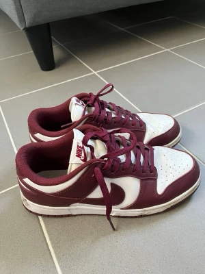 Nike Dunk Low Bordeaux sneakers - Snygga Nike Dunk Low sneakers i färgen bordeaux och vitt. Skorna har klassisk låg siluett, perforerad tåbox och matchande bordeauxfärgade skosnören. Ovandelen är i läder med vit bas och bordeaux detaljer, samt Nike-logga på plösen och sidan. Yttersulan är också bordeaux.