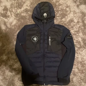 Mörkblå Sail Racing pufferjacka - Snygg mörkblå pufferjacka från Sail Racing med huva och dragkedja. Jackan har broderad Sail Antarctica Expedition-patch på bröstet och logga på bröst och huva. Tillverkad i polyester med quiltade partier och långa ärmar. Perfekt för kyliga dagar. Använd bara en vinter.