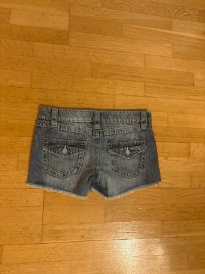 Blå lågmidjade jeansshorts med slitningar - Säljer ett par blå lågmidjade jeansshorts med råa kanter och slitna detaljer. Shortsen har fem fickor, bälteshällor och knappstängning fram. Perfekta för varma dagar och en avslappnad stil. Storleken är 158 men passar xs. Priset går att diskutera. Frakt ingår ej! Använd en gång.