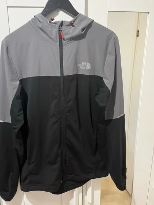 Svart och grå vindjacka The North Face - Snygg vindjacka från The North Face i svart och grått med dragkedja framtill och huva. Jackan har logga på bröstet och röda detaljer vid snörningen. Perfekt för blåsiga dagar och enkel att matcha med olika outfits.