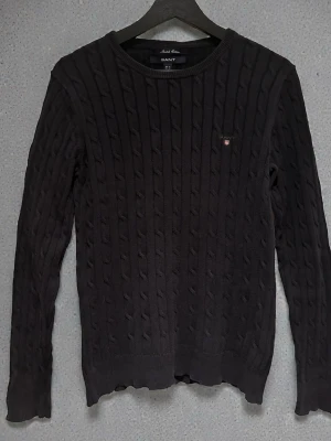 Svart kabelstickad GANT tröja  - Kabelstickad svart tröja från GANT med rund halsringning och diskret broderad logga på bröstet. Tröjan är i nyskick, och inga defekter!                 Storlek: M