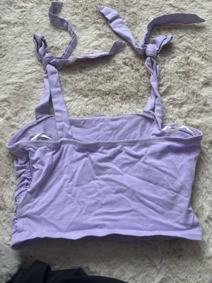 Ljuslila linnetopp med knytband - Supersöt ljuslila linnetopp med knytbara axelband och rynkade sidor. Toppen är ärmlös och har en croppad passform som passar perfekt till höga jeans eller kjol. Tillverkad i mjuk bomull för en bekväm känsla.