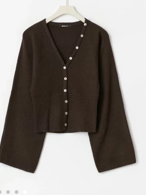 Brun kofta med vida ärmar från Gina Tricot - Mörkbrun stickad kofta från Gina Tricot med v-ringning och dekorativa vita knappar längs hela framsidan. Koftan har vida ärmar och ribbad midja som ger en snygg siluett. Perfekt för lager på lager och en trendig look. Har prislappen kvar!!
