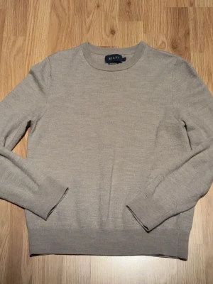 Beige stickad tröja från Riley - Säljer en stilren beige stickad tröja från Riley i mjuk merinoull. Tröjan har rund halsringning, långa ärmar och ribbade muddar vid ärmslut och nederkant. Perfekt för lager på lager och enkel att matcha med olika outfits.