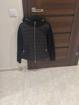 Svart quiltad pufferjacka med huva - Snygg svart pufferjacka med quiltad front, dragkedja och huva med snörning. Ärmar och baksida är i mjukare material för en cool kontrast. Perfekt för dig som vill ha en stilren jacka med sportig touch.