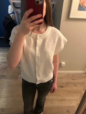 Vit kofta med knappar - Säljer en söt vit kofta med korta ärmar och rund hals. Koftan har ribbstickade kanter och knäpps med vita knappar framtill. Perfekt att slänga över en topp för en clean och enkel look.