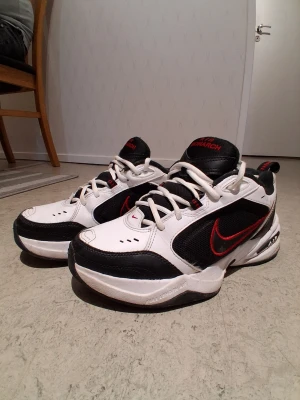 Nike Air Monarch vita och svarta sneakers - Nike Air Monarch sneakers i vitt och svart med röda detaljer och logga. Skorna har en chunky sula, meshpaneler för ventilation och klassisk snörning. Perfekta för dig som gillar retrostil och bekväma sneakers med sportig känsla.