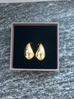 Guldiga droppformade örhängen - Säljer ett par snygga örhängen i droppformad design med blank guldfinish. De är chunky och har en modern, stilren look som verkligen sticker ut. Perfekta för dig som gillar statement-smycken och vill addera lite extra bling till din outfit.