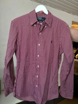 Rödvit randig Ralph Lauren skjorta - Klassisk röd och vit randig skjorta från Polo Ralph Lauren med button-down krage. Klassiska loggan på bröstet och vita knappar framtill. Slim fit men luftigt material så den sitter skönt på.