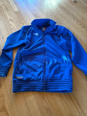 Blå träningsjacka från Diadora stl 170 - Snygg blå träningsjacka från Diadora med hel dragkedja och ståkrage. Jackan har ribbade muddar och nederkant med svarta ränder samt en vit logga på bröstet. Tillverkad i ett mjukt och funktionellt material, perfekt för sport och träning.