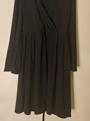 Svart omlottklänning med lång ärm - En svart kortklänning med omlottliv och långa ärmar. Klänningen har en lätt utställd kjol och markerad midja, vilket ger en smickrande siluett. Perfekt för dig som gillar enkel och stilren design.