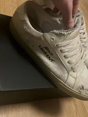 Saint Laurent vita låga sneakers - Vita sneakers från Saint Laurent med handskriven logga på sidan. Skorna har slitna detaljer, vita snören och klassisk låg profil. Tillverkade i canvas och läder för en cool och avslappnad look.