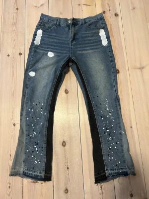 Blå jeans med färgstänk och svarta detaljer - Unika blå jeans med vita färgstänk och svarta paneler längs benen. Jeansen har raka ben, vita detaljer vid fickorna och coola konstnärliga splash-effekter framtill. Perfekta för dig som vill sticka ut med en kreativ och trendig look.