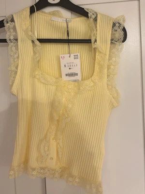Gul ribbad spetslinnetopp - Supersöt ljusgul ribbad linnetopp från Zara med spetsdetaljer runt halsringning, ärmhål och nederkant. Toppen har knappar framtill och är ärmlös, perfekt för en somrig och feminin look. Prislapp kvar!