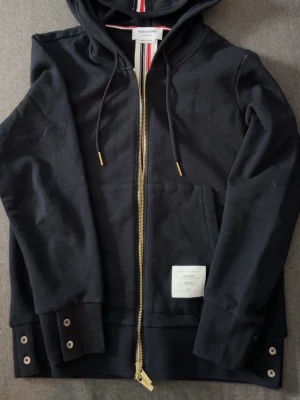 Mörk blå zip hoodie från Thom Browne - Säljer en svart hoodie från Thom Browne med dragkedja i guld, huva med snören och ribbade muddar. Tricolore-detalj i luvan och knappdetaljer vid ärmslut och nederkant. Tillverkad i mjuk bomull och har en vit patch med logga på framsidan. Det är storlek 1 vilket mot svarar Medium