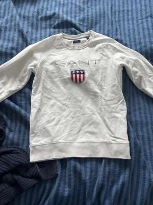 Vit sweatshirt från GANT med logga - Vit sweatshirt från GANT med broderad logga och sköld med stjärnor och ränder på bröstet. Tröjan har rund halsringning och långa ärmar. Perfekt för en clean och sportig stil.