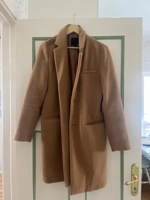 Kamelbrun kappa från 157 - Stilren kamelbrun kappa från 157 med klassisk krage och tre fickor framtill. Kappan har rak passform och är helfodrad för extra komfort. Perfekt för dig som gillar en tidlös och clean look. Materialet känns mjukt och ger en exklusiv känsla.