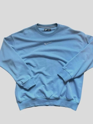 Blå sweatshirt från Nike - Snygg ljusblå sweatshirt från Nike med klassisk rund halsringning och broderad vit Nike-logga på bröstet. Tröjan har ribbade muddar vid ärmslut och nederkant, perfekt för en avslappnad och sportig stil.