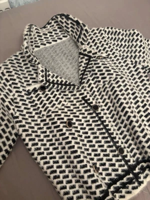 Svartvit mönstrad cardiganjacka - Säljer en svartvit cardiganjacka med grafiskt mönster och dekorativa svarta knappar med guldkant. Jackan har krage, dubbelknäppning och svarta kantband. Materialet är mjukt och ser ut att vara ullblandning. Perfekt för dig som gillar en trendig och unik look.