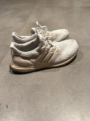Adidas Ultra Boost sneakers - Snygga Adidas Ultra Boost sneakers i beige och vitt med stickad ovandel och klassiska tre ränder på sidan. Skorna har en bekväm Boost-sula och snörning framtill. Perfekta för dig som gillar stilrena och sportiga sneakers.