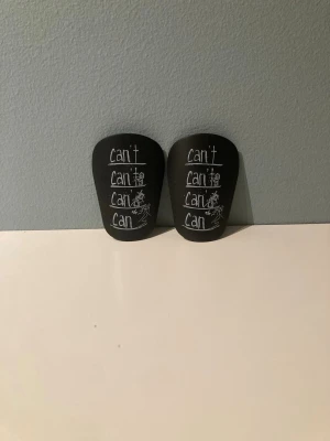 Svarta benskydd med text - Säljer ett par svarta benskydd med vit text där det står 'can't' flera gånger och 'can' längst ner, samt en tecknad figur. Perfekta för fotboll eller annan sport där du behöver extra skydd för benen. Lätta och smidiga att använda under träning eller match.