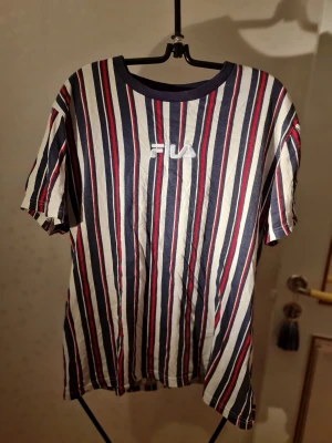 Randig t-shirt från Fila - Säljer en randig t-shirt från Fila med marinblå, röda och vita ränder. T-shirten har rund halsringning och korta ärmar. Tillverkad i mjuk bomull som känns skön mot huden. Perfekt för en avslappnad och sportig stil.