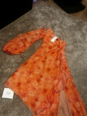 Orange blommig oneshoulder klänning - Säljer en unik orange oneshoulder klänning från Adoore med blommigt mönster och volangdetalj framtill. Klänningen har en lång puffärm och är tillverkad i ett lätt, transparent material som ger en luftig känsla. Perfekt för dig som vill sticka ut!