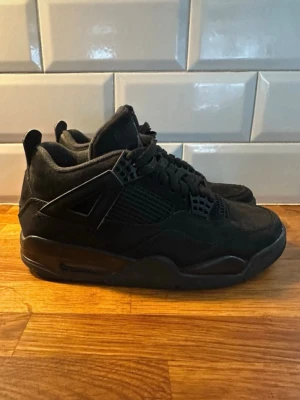 Svarta Air Jordan 4 Retro sneakers - Snygga helsvarta Air Jordan 4 Retro sneakers i mocka och mesh. Klassisk siluett med coola detaljer som plastvingar och Jumpman-logga på plösen. Perfekta för dig som gillar streetwear och vill ha en stilren look.