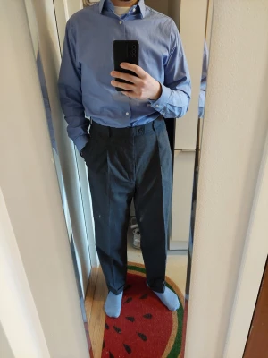 Kostymbyxor Stuart Hughes mörkgrå - Kostymbyxor med klassisk passform med högre midja och veck på framsidan. Mycket bekväma i 100% mörkgrå ull, smått rutigt mönster i tyget. Woolmark märkt. Storlek W34L32.