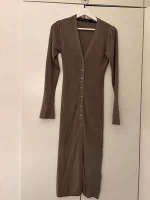 Beige/brun långklänning/kofta med knappar - Långklänning i beige/brun ribbat med v-ringning och långa ärmar. Klänningen har knappar hela vägen framtill och en smickrande, figurnära passform. Perfekt för dig som gillar stilrena och bekväma plagg. Fungerar även som kofta. Xs men passar även S.
