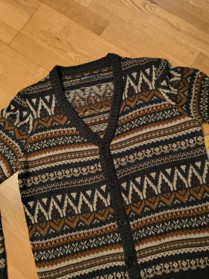 Mönstrad kofta  - Snygg kofta med knappar i mörkgrått, brunt, beige och vitt. Koftan har ett klassiskt nordiskt mönster, v-ringad hals och ribbade muddar. Perfekt för dig som gillar retrovibbar och stickade plagg.