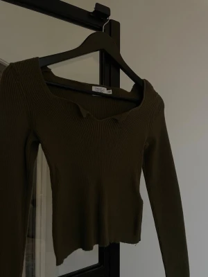 Olivgrön ribbad  topp - Trendig olivgrön  topp från nakd med lång ärm och ribbad struktur. Toppen har en unik v-ringad halsringning med vågig kant som ger en cool detalj. Perfekt att styla med höga jeans eller kjol (sticks ej på huden ) . Är xxs men passar mer som xs skulle jag säga 
