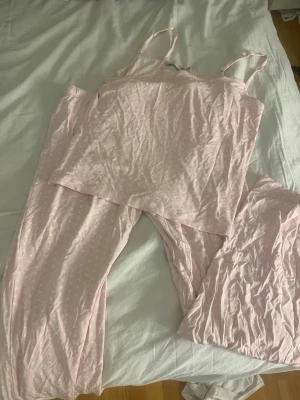 Ljusrosa pyjamas-set med linne - Säljer ett ljusrosa pyjamas-set med linne och långa byxor. Linnet har smala axelband och byxorna är vida med resår i midjan. Materialet är mjukt och tunt, perfekt för att chilla hemma. Diskret prickigt mönster på byxorna. Supermysigt set för kvällar och morgnar!