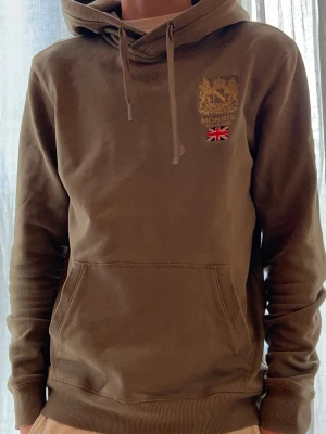 Olivgrön hoodie från Morris ST s sitter som xs - Olivgrön hoodie från Morris med broderad logga och brittisk flagga på bröstet. Klassisk känguruficka, huva med dragsko och ribbade muddar. Perfekt för en avslappnad och stilren look. Sitter mindre än storleken 