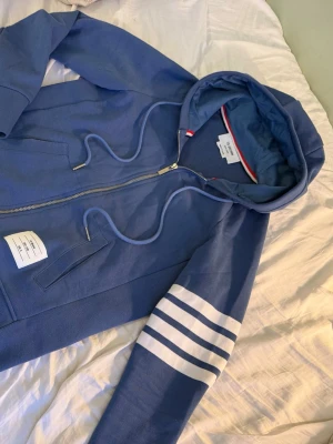Thom Browne hoodie - Snygg blå hoodie från Thom Browne med dragkedja, vita ränder på ena ärmen och klassiska detaljer som snörning i huvan och fickor framtill. Märkeslapp på magen och diskreta röd-vita detaljer i halsen. Perfekt för en chill och stilren look.