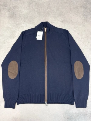 Marinblå ziptröja Gran Sasso - Stilren marinblå ziptröja från Gran Sasso med ribbad ståkrage och kontrasterande bruna mockadetaljer på armbågarna. Tröjan har hel dragkedja framtill och ribbade muddar vid ärmslut och nederkant. Perfekt för en snygg och avslappnad look.