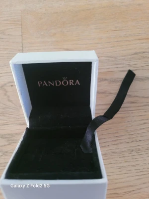 Vit smyckesask från Pandora - Snygg ask från Pandora med svart inredning och ett svart band. Perfekt för att förvara eller ge bort smycken. Locket har Pandoras logga i silvertext och asken har en stilren, fyrkantig form.