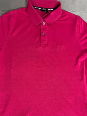 Hugo Boss pikétröja   - Säljer en snygg rosa pikétröja från Hugo Boss med klassisk krage och två knappar framtill. Tröjan har diskret broderad logga på bröstet och är tillverkad i ett mjukt bomullsmaterial. Perfekt för dig som gillar stilrena och färgstarka plagg.