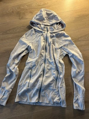 Ljusgrå hoodie från Lululemon - Snygg ljusgrå hoodie från Lululemon med dragkedja framtill och två fickor med dragkedja. Tillverkad i mjukt och stretchigt material, perfekt för chill eller träning. Har en stor huva och tumhål vid ärmsluten. Lululemon-logga på ärmen.