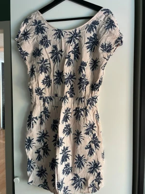 Beige kortklänning med blå palmer - Kortklänning i beige med marinblått palmtryck, tillverkad i luftig bomull. Klänningen har korta ärmar, resår i midjan och en öppen detalj i ryggen som ger en chill vibe. Perfekt för sommardagar när du vill vara både bekväm och snygg.