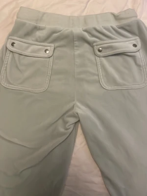 Ljusgrå velourbyxor Juicy Couture - Säljer ett par ljusgrå mjukisbyxor från Juicy Couture i mjuk velour. Byxorna har bred resår i midjan, dragsko med metalländar och stora fickor både fram och bak med lock och tryckknappar. Klassisk JC-logga broderad framtill.